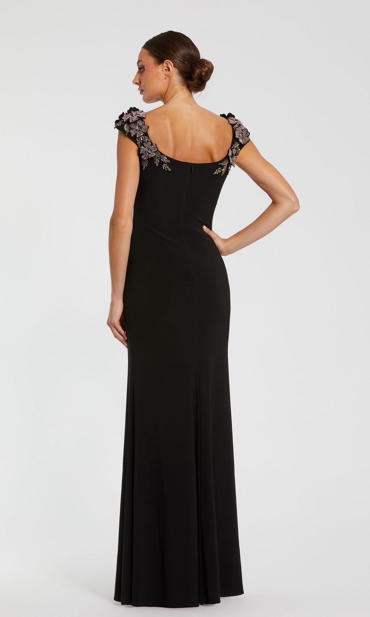 Woman in a Mac Duggal 11842 long formal dress with 3D floral embellishments and cap sleeves, standing with her back to the camera, hair in a low bun, set against a plain background.